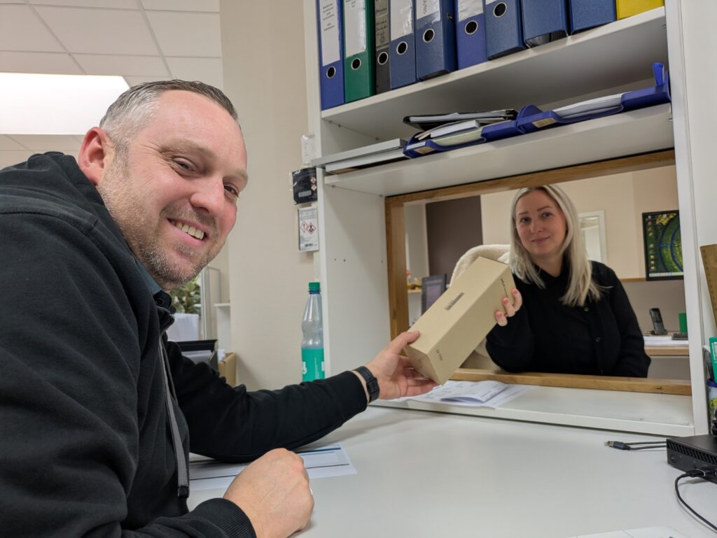 Mitarbeiterin überreicht einem Kollegen ein Paket an einem Büro-Schalter, beide lächeln freundlich.