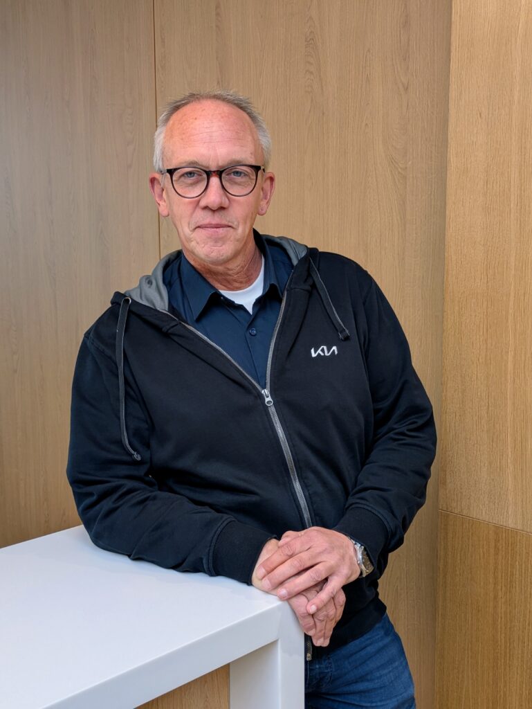 Mitarbeiter im KIA-Servicebereich steht lächelnd mit Brille an einem weißen Tresen vor Holzrückwand.