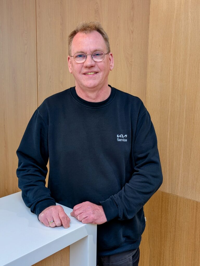 Mitarbeiter im KIA-Servicebereich steht lächelnd an einem weißen Tresen vor Holzrückwand.