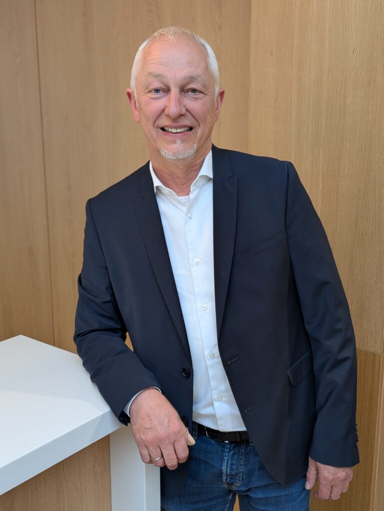 Mitarbeiter Dirk Masemann im KIA-Servicebereich steht lächelnd an einem weißen Tresen vor Holzrückwand.