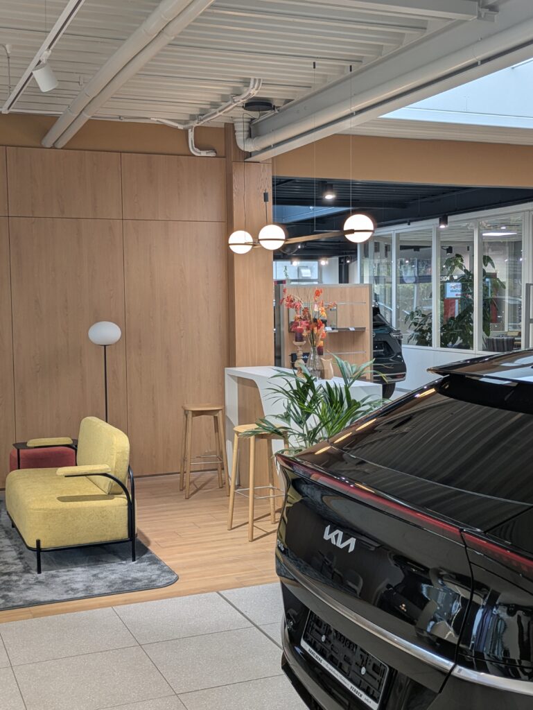 Moderner KIA-Ausstellungsraum mit Sitzbereich, Holzwand, Pflanzen und Fahrzeug im Vordergrund.