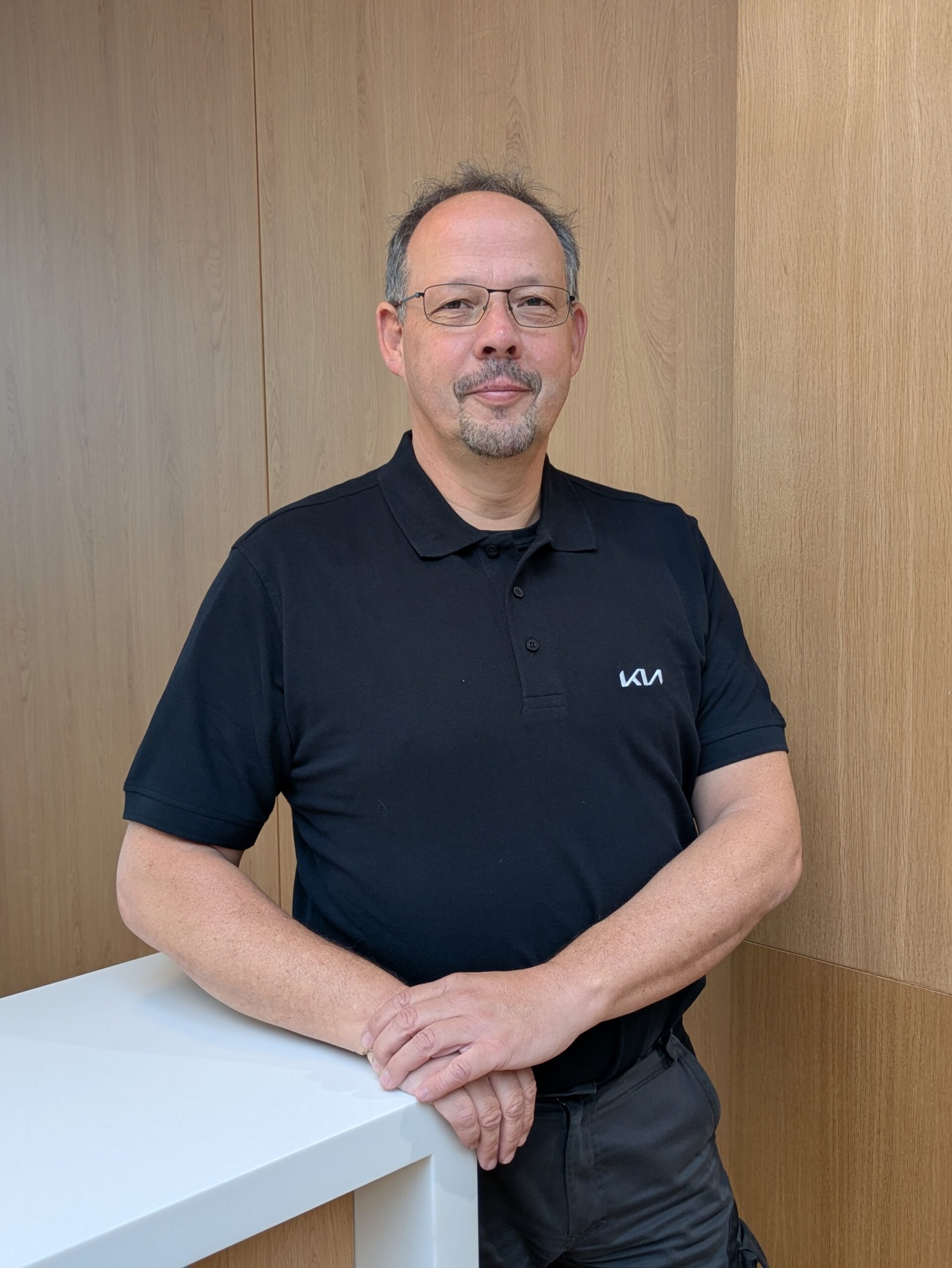 Mitarbeiter mit Brille im KIA-Servicebereich steht lächelnd an einem weißen Tresen vor Holzrückwand.